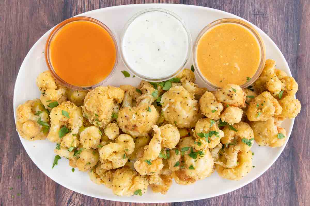 Deep fried cauliflower bites on a white platter with three dipping sauces.