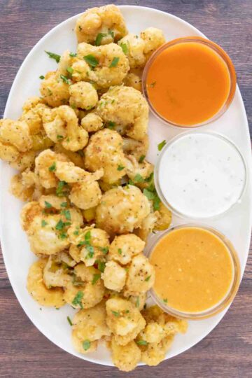 Deep fried cauliflower bites on a white platter with three dipping sauces.