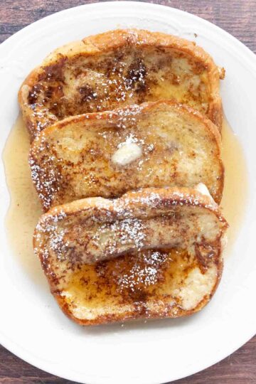 French Toast with butter, maple syrup and confectioners sugar on a white plate.