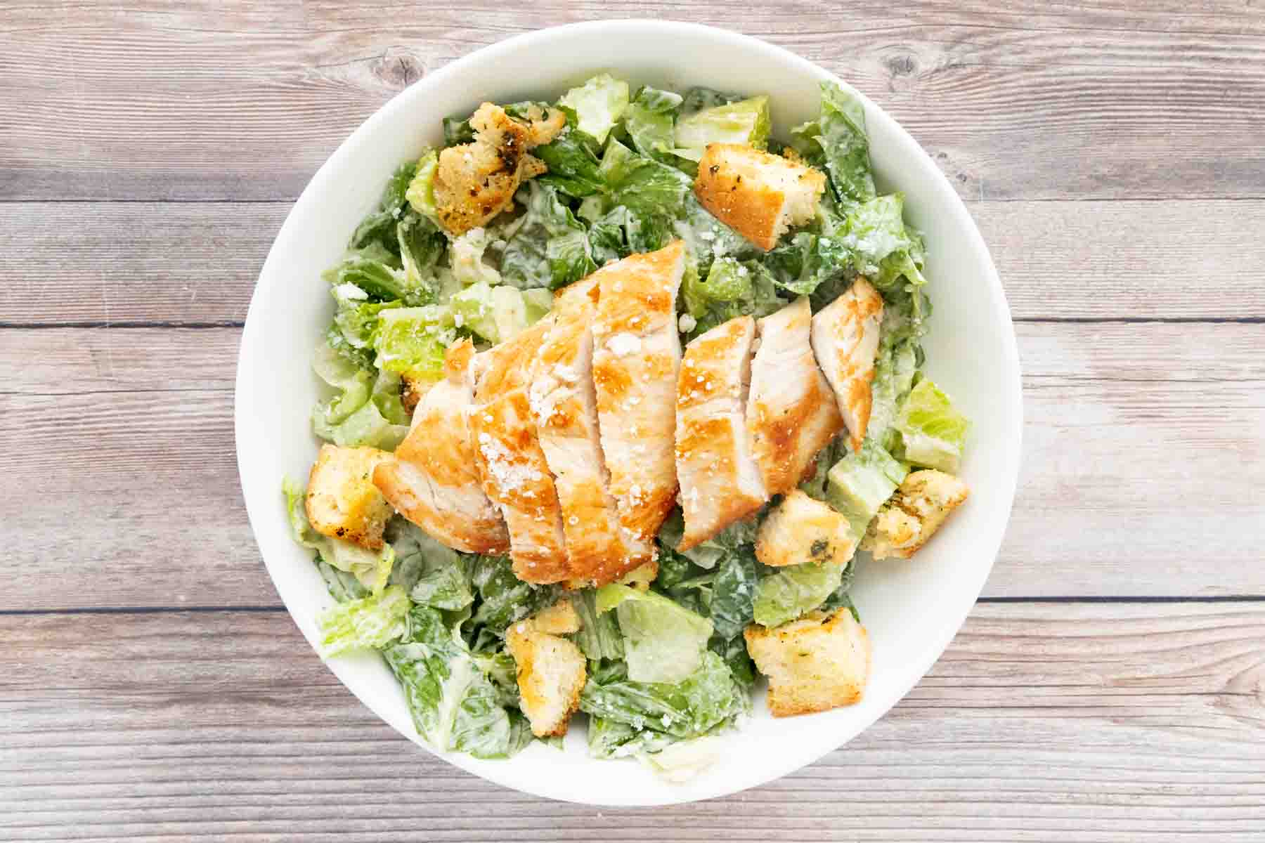 Bowl of Caesar salad with croutons topped with grilled chicken.