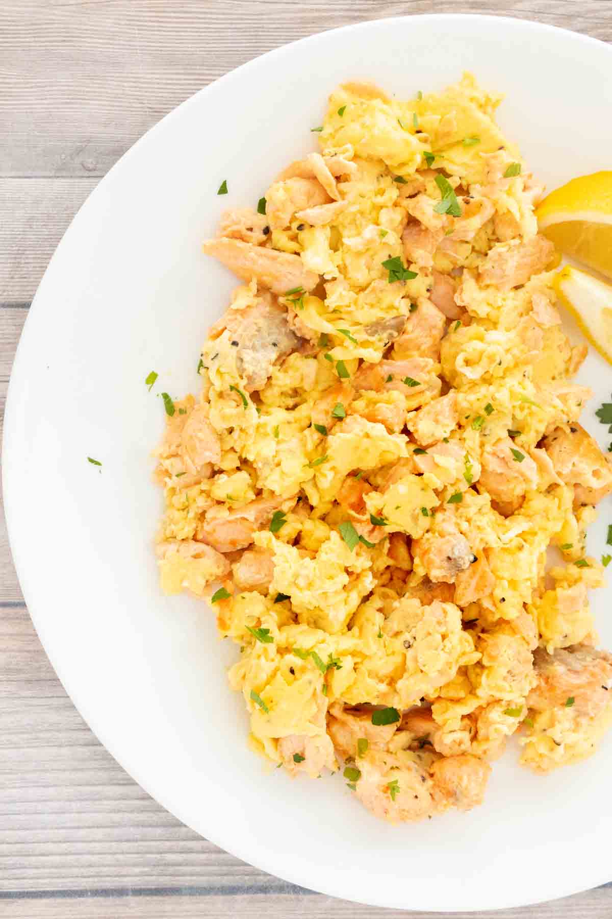 A white plate with a mixture of scrambled eggs and salmon. Garnished with fresh parsley and lemon wedges.