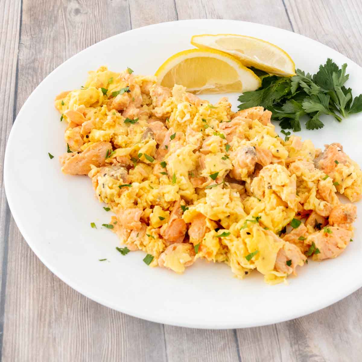 A white plate with a mixture of scrambled eggs and salmon. Garnished with fresh parsley and lemon wedges.